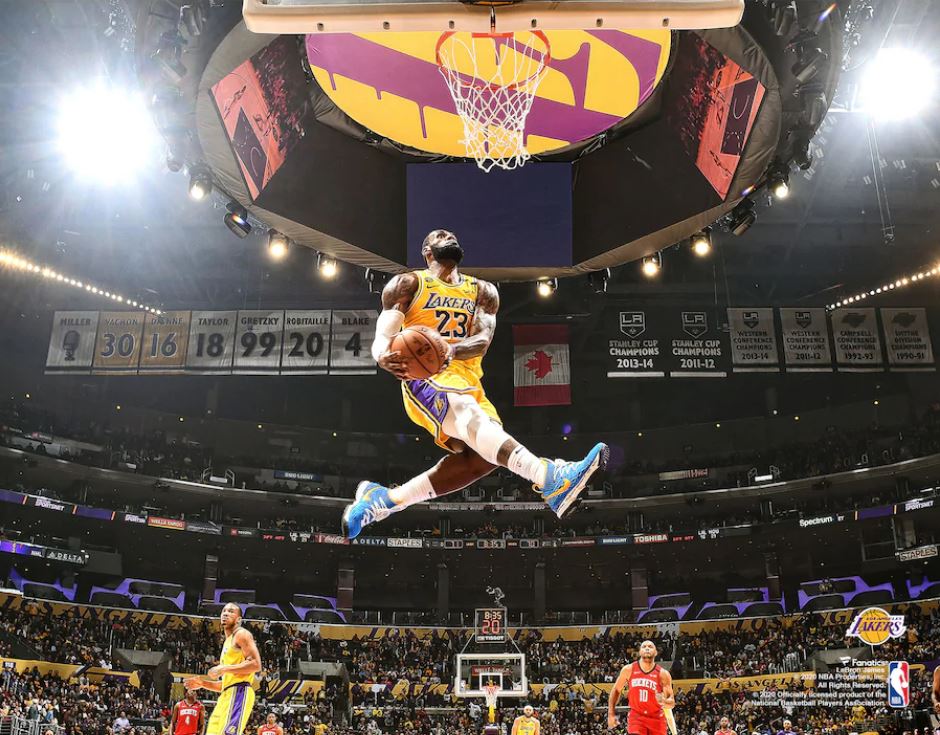 A dynamic action shot of LeBron James wearing Lakers jersey #23 in yellow and purple, captured mid-dunk at Staples Center (now Crypto.com Arena). The image shows James in athletic flight with the basketball, demonstrating his athleticism and skill. The arena is fully lit with crowd visible in the background, retired jersey numbers displayed on the upper level, and Stanley Cup championship banners visible. The mood is energetic and celebratory, capturing an iconic moment during an NBA game. The photograph is taken from a ground-level perspective looking up, emphasizing the height and power of the dunk.