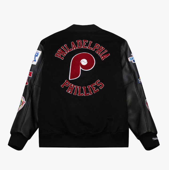 Black Philadelphia Phillies varsity jacket shown from the back, featuring a large red and white Phillies 'P' logo with 'PHILADELPHIA PHILLIES' text embroidered in red and white. The sleeves are made of black faux leather, and several team patches are visible on the sleeves.