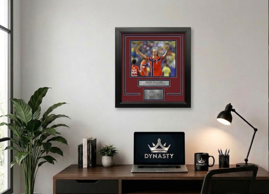 A framed sports memorabilia piece featuring a photo of Erling Haaland in an red and blue Norway national football team jersey, with a red mat, black frame, and a silver plaque at the bottom containing a replica signature. The photo is hanging in an office.