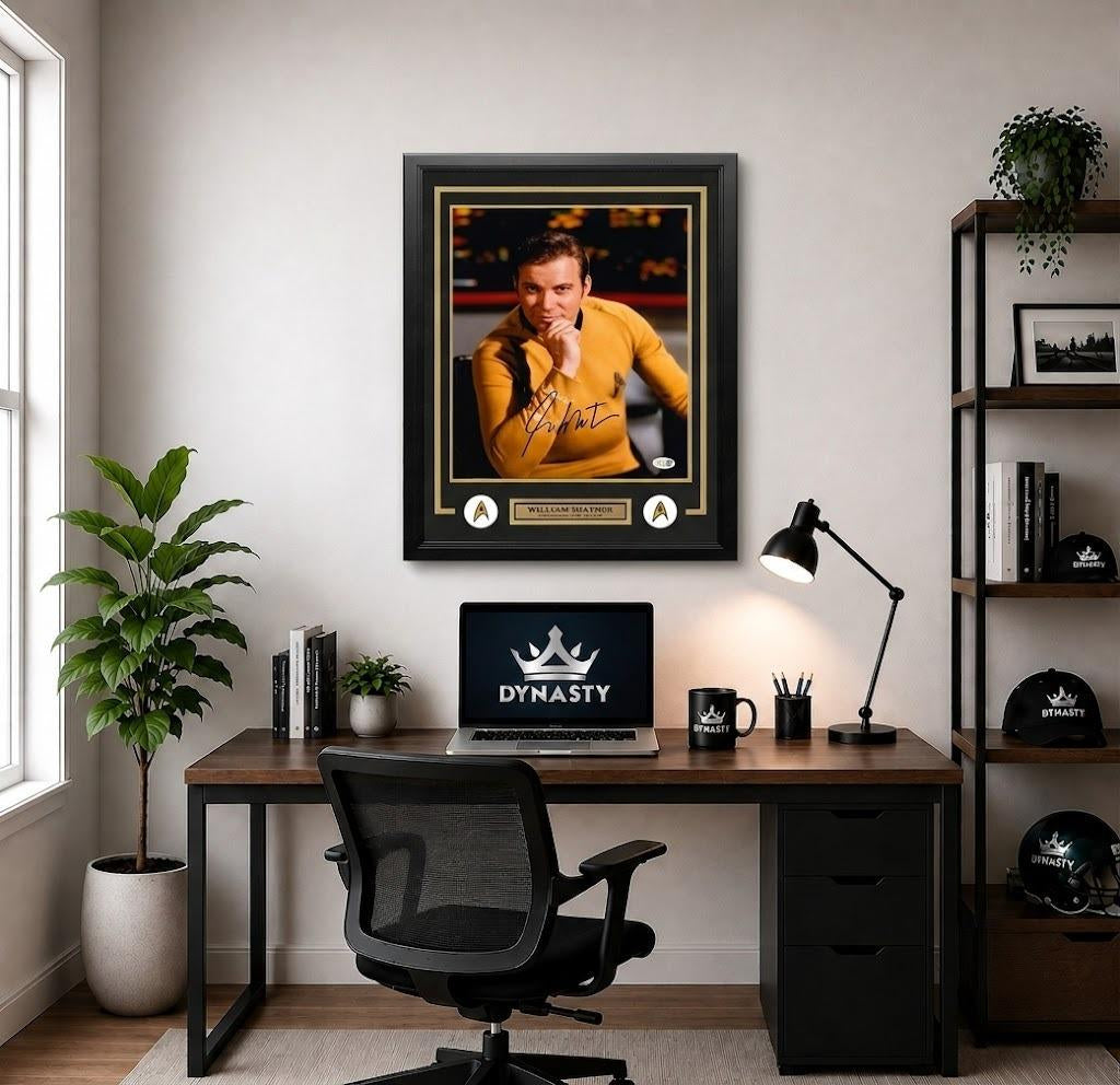 A framed autographed sports photo of Williams Shatner as Captain Kirk in a displayed on a dark wood desk in a modern home office with a laptop, lamp, and plants.
