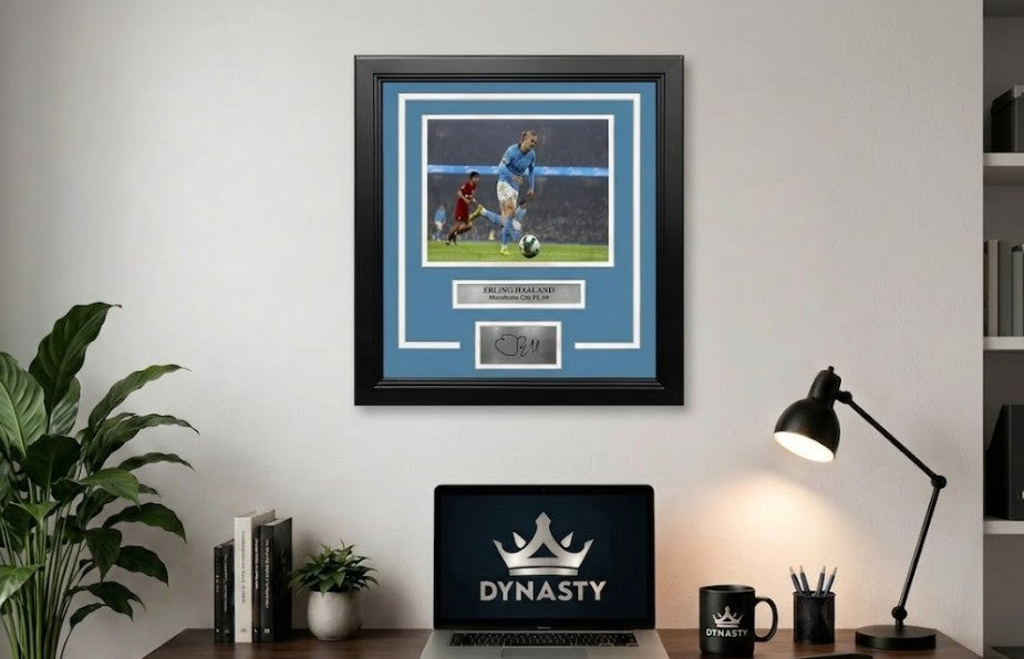 A framed sports memorabilia piece featuring a photo of Erling Haaland in an white and blue Manchester City FC team jersey, with a sky blue mat, black frame, and a silver plaque at the bottom containing a replica signature. It is hanging in an office