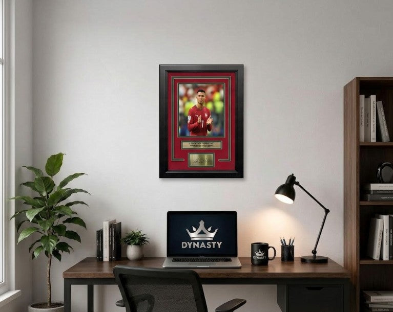 Framed Cristiano Ronaldo photo featuring a portrait of the player in a Portugal national team jersey, with a red mat, green inner border, black frame, and gold-toned nameplate and signature plaque. It shows Ronaldo celebrating his final World Cup match in 2026. It is hanging in an office.