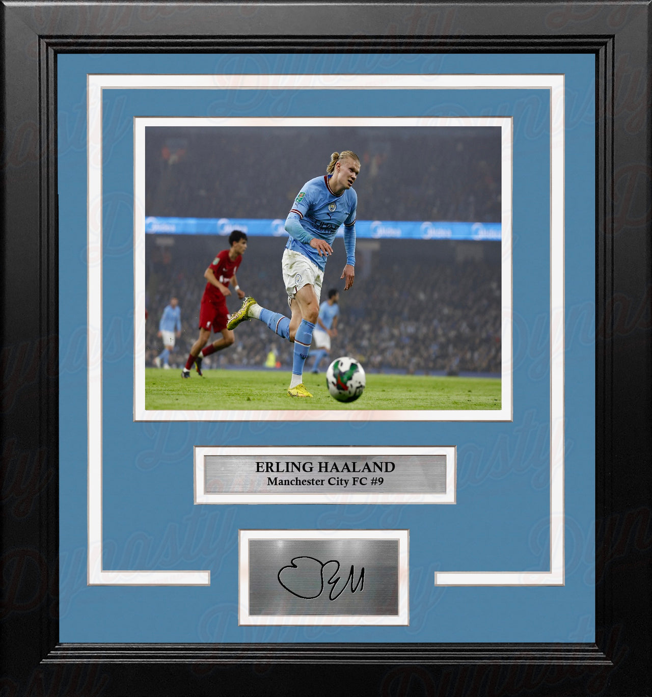 A framed sports memorabilia piece featuring a photo of Erling Haaland in an white and blue Manchester City FC team jersey, with a sky blue mat, black frame, and a silver plaque at the bottom containing a replica signature.