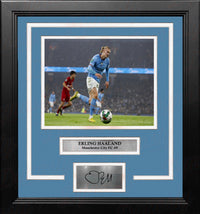 A framed sports memorabilia piece featuring a photo of Erling Haaland in an white and blue Manchester City FC team jersey, with a sky blue mat, black frame, and a silver plaque at the bottom containing a replica signature.