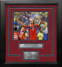 A framed sports memorabilia piece featuring a photo of Erling Haaland in an red and blue Norway national football team jersey, with a red mat, black frame, and a silver plaque at the bottom containing a replica signature.